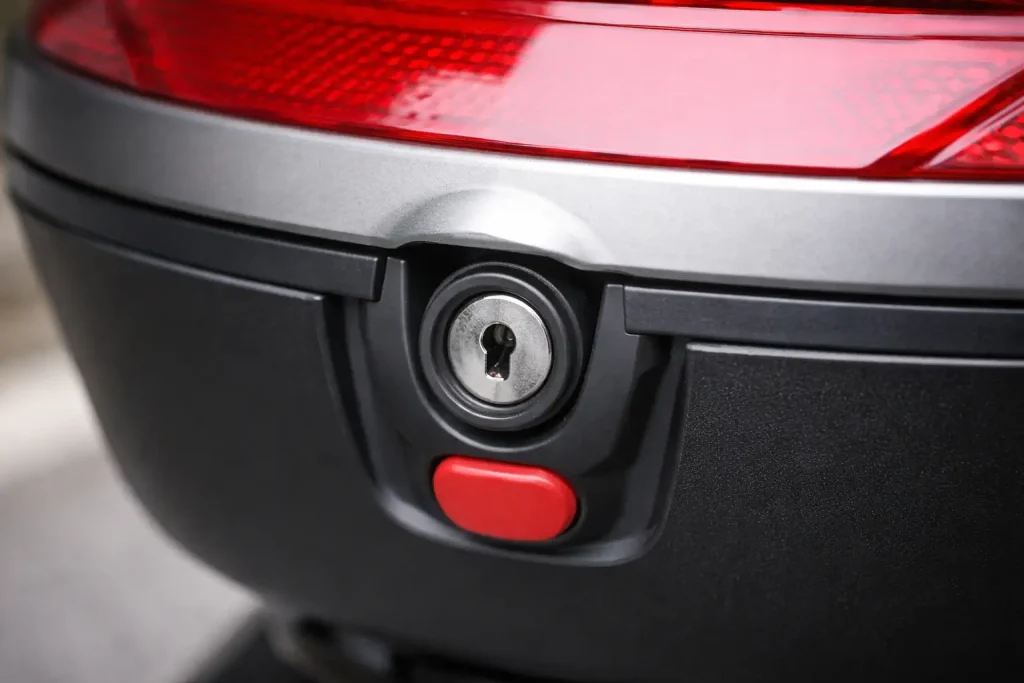 Gros plan sur la serrure d’un top-case moto sans clé insérée, avec bouton d’ouverture rouge sous le barillet
