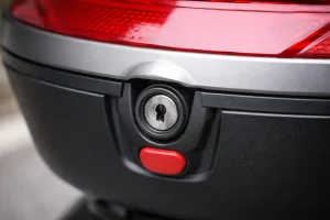 Gros plan sur la serrure d’un top-case moto sans clé insérée, avec bouton d’ouverture rouge sous le barillet